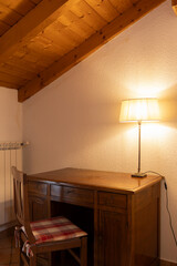 Detail of a corner of a bedroom with a desk a chair and a lamp with a light on. Everything looks old