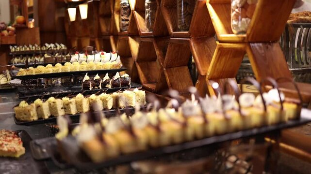 Elegant dessert buffet lined with rows of colorful mini cakes topped with cream and chocolate curls set against warm wooden shelves and glass jars in a luxurious sweet display