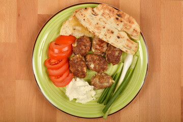An overhead shot shows a green plate featuring grilled savory minced meat patties with cheese, sliced tomatoes, spring onions, pita bread, and a dollop of white kajmak on wooden surface