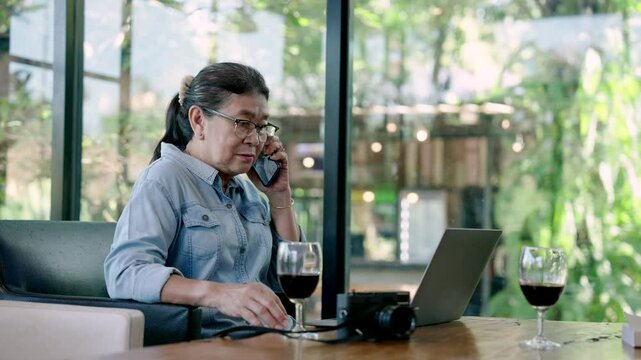 asian senior woman talks on phone while working on laptop at cafe during retirement trip surrounded by wine glass camera wooden table modern natural light indoor scenery