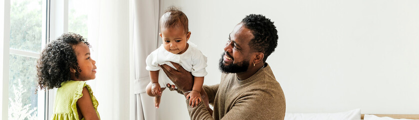 Joyful Black father holding baby while talking with his daughter near a window in a cozy bedroom. A warm, diverse family moment celebrating fatherhood, love, and authentic everyday parenting.