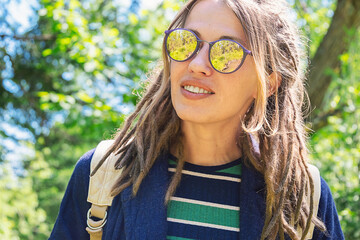 Woman with Dreadlocks and Sunglasses Stands Outdoors in a Woods, Enjoying a Sunny Day.