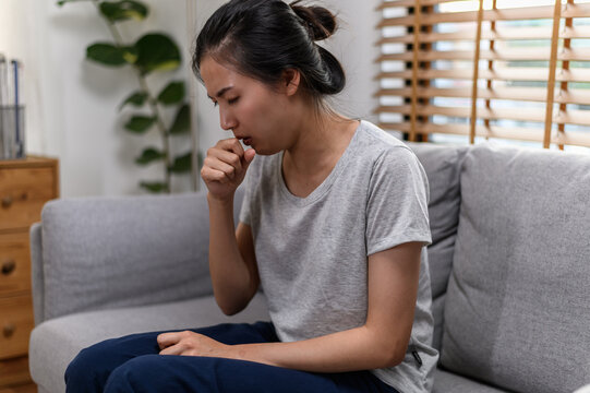 Asian woman sitting on sofa and coughing, showing symptoms of illness. Concept of cold, flu, respiratory infection, or seasonal sickness. Healthcare and wellness at home.