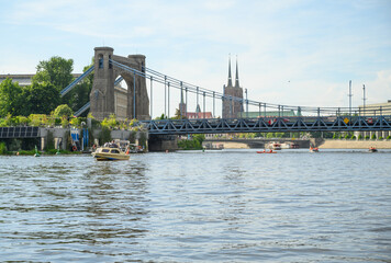 Obraz premium Urban river scenery featuring the Grunwaldzki suspension bridge and neo-Gothic cathedral on a clear afternoon in Wroclaw, Poland.