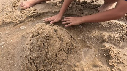 A child is building a sandcastle with their hands near a small pool of water on the beach.
