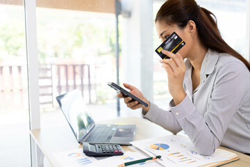 A woman is sitting at a desk with a laptop and a cell phone. She is looking at her cell phone and...