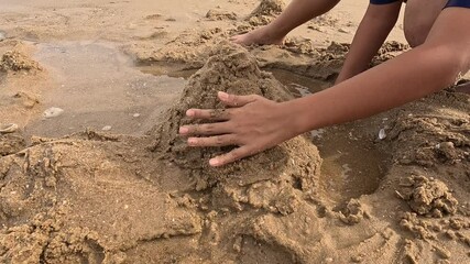 A child is building a sandcastle with their hands near a small pool of water on the beach.
