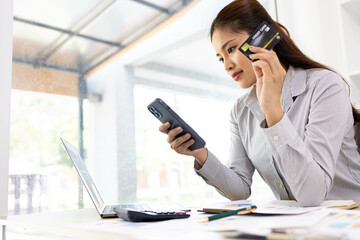 A woman is sitting at a desk with a laptop and a cell phone. She is looking at her cell phone and...