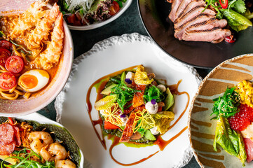 A vibrant selection of gourmet dishes including ramen with tempura shrimp, fresh salad with avocado and citrus, seared duck breast, and a seafood bowl with shrimp, seaweed, and corn, beautifully prese