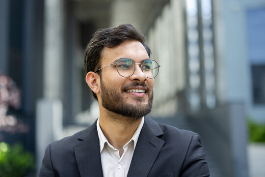 A smiling man in a suit and glasses poses outdoors, looking thoughtful and confident in a city setting. - Powered by Adobe