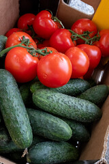 A close-up, top-down view of a cardboard box filled with vibrant red vine-ripened tomatoes and fresh green cucumbers, showcasing healthy, natural produce from a market.