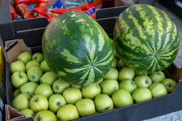 Two large, ripe watermelons displayed on top of a box full of crisp green apples at a vibrant outdoor fruit market