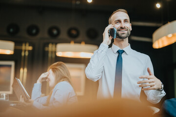 Smiling businessman in a white shirt talking on a phone indoors, with coworkers in a modern office...