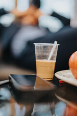 A plastic cup of coffee with a straw placed near a smart phone on a reflective table, with a blurred background suggesting a relaxed or casual office setting.