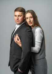 Stylish man in suit stands half turned, young woman embraces him from behind. They look at camera. Studio portrait of stylish couple. Concept of love, trust and style