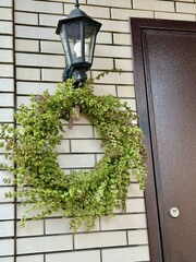 decorative wreath of fresh twigs, sage flowers and leaves with a purple ribbon hangs against the background of a light cypress wall next to the front door to the house.