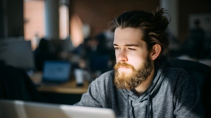 Obraz premium Young Man with Beard Focused on Laptop in Bright Workspace Environment, Deep in Thought