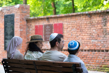 Jewish family sits on bench in front of memorial wall in memory of those killed in Holocaust, mother, father, son and daughter mourn near monument on common Jewish grave