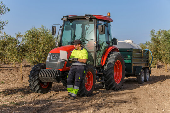 Olive grower operating a laptop computer sitting on the wheel of a tractor trailer in an Andalusian olive grove. - Powered by Adobe