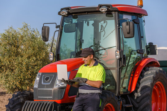 Farmer using laptop near red tractor in olive grove