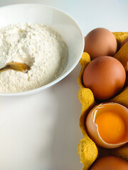 Minimalist baking scene with flour, yellow spoon, brown eggs in yellow carton, and one cracked egg on white background.