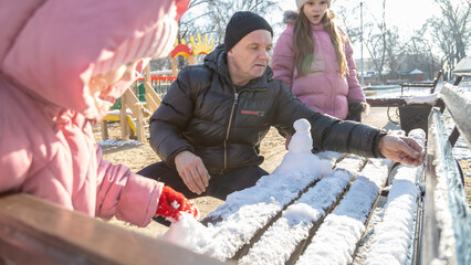 Grandfather with granddaughters making a little snowman in sunny winter day