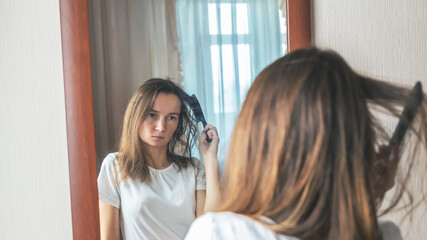 Fototapeta premium Young woman brushing hair brush looking at the mirror at bedroom