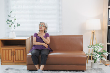 Fototapeta premium Elderly asian woman suffering from chest pain, having heart attack or breathing difficulties, sitting on sofa at home, senior healthcare and medical concept