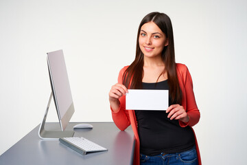 Smiling white adult woman in red jacket holding blank sign while standing at office desk with computer. Concept of business promotion, marketing, and corporate communication