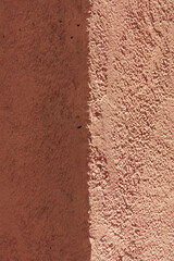 Two distinct stucco wall surfaces meet. One side shaded and dark brown. The other brightly illuminated, showing clay earth tones and textures. Strong visual contrast between two sides of building.