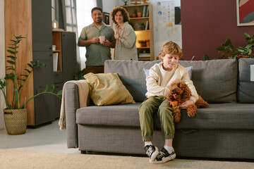 Obraz premium Caucasian child sitting on sofa petting small brown dog while smiling, with Caucasian middle aged man and woman standing together in background watching child interact with first pet at home