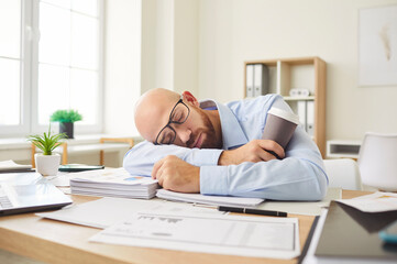 Tired bald young man wearing glasses sleeping at his desk full of documents in bright office. Exhausted overworked employee taking nap, resting his head on his arms, holding disposable coffee cup.