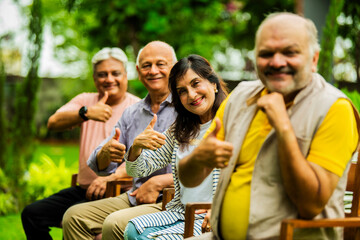 Indian seniors pointing and smiling together, seated outdoors in green garden with thumbs up
