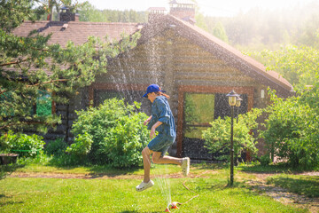 Happy girl with blue cap and denim dress jumping and playing with water jets from a sprinkler in the garden of a country house during a sunny summer day