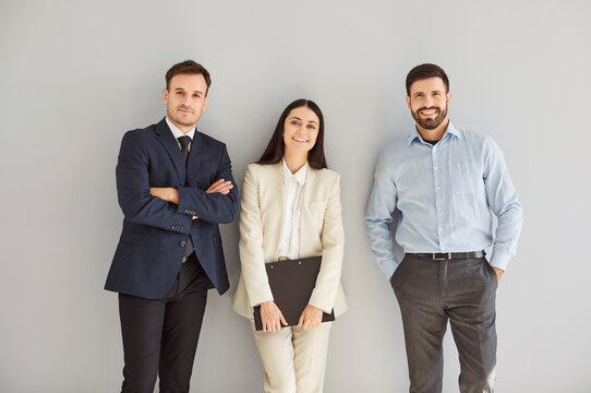 Portrait of a group of three young cheerful business people in suits standing confidently, looking at the camera and smiling isolated on a studio grey background. Team work concept.
