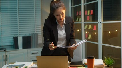 Business Asian woman in formal black suit thoughtfully reviews documents at her desk late at night, critical thinking, and commitment to meeting deadlines during late-night work hours.