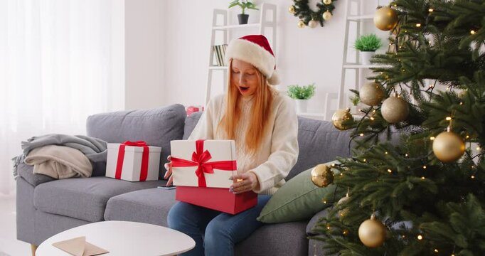 Young woman receives Christmas surprise. Happy girl in white sweater and red Santa Claus hat on sofa by decorated tree in living room opens magic gift box and takes out small box with jewellery inside