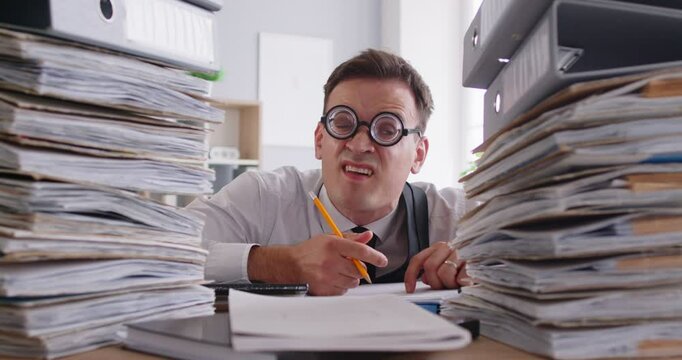 Funny busy focused hard working man bookkeeper, office worker, clerk in thick glasses sits at desk, sticks tongue out, diligently writes notes and looks at paperwork piles with comical face expression