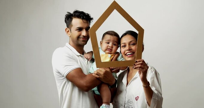 Indian young parents and baby boy looking through house shaped cardboard cutout to promote real estate investment, isolated on white background, representing happy Asian family and housing concept