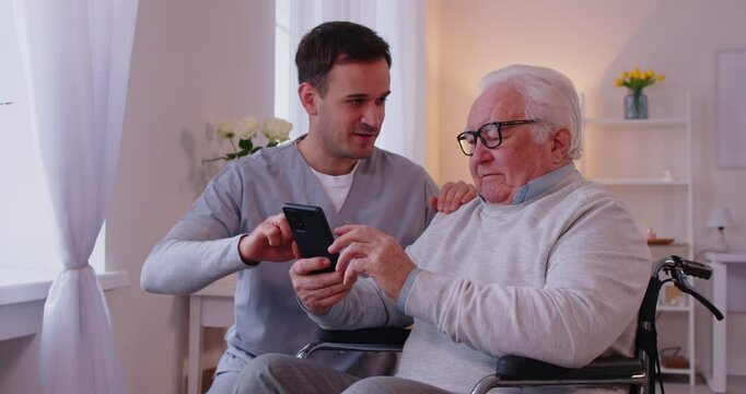 Male caregiver or social worker teaching senior man with disability in wheelchair how to use mobile phone. Nurse learning his elderly disabled patient using smartphone in nursing home. 4k video.