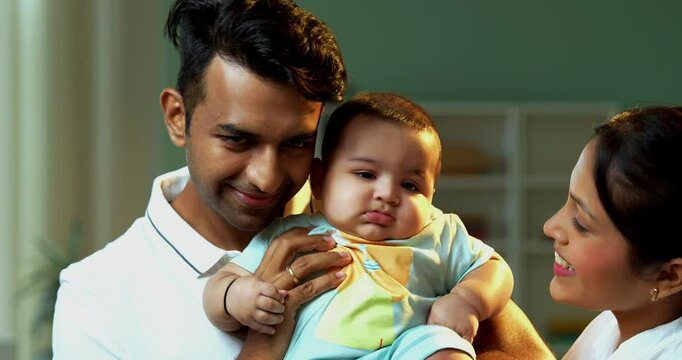 Indian parents kissing baby boy on chicks while standing together in cozy living room, smiling with love, warmth, expressing emotional bonding, happiness, protection, and parenthood at home indoors