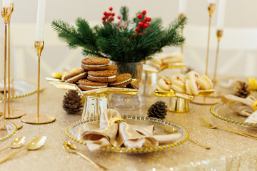 Luxury Christmas dinner table setting. Fir branches in vase, chocolate cookies, macaroons, golden napkin on glass plate, cutlery, candlesticks on tablecloth. Xmas holidays. Festive new 2026 year party