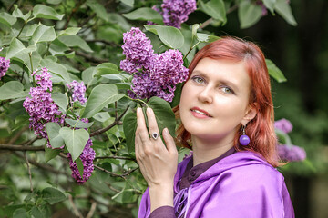 A beautiful young woman with red hair, against a background of blooming lilacs, in a cloak and...