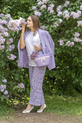 A beautiful young blonde woman, against a background of blooming lilacs, in a lilac trouser suit. Femininity and nature.