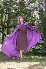 A beautiful young woman with red hair, against a background of blooming lilacs, in a cloak and hood. Medieval fantasy costume. Fairy tale heroine, princess, duchess.