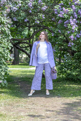 A beautiful young blonde woman, against a background of blooming lilacs, in a lilac trouser suit. Femininity and nature.