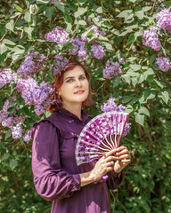 A beautiful young woman with red hair, against a background of blooming lilacs, in a cloak and hood. Medieval fantasy costume. Fairy tale heroine, princess, duchess.