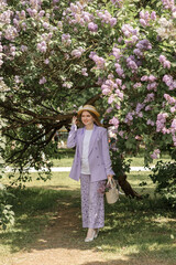 A beautiful young blonde woman, against a background of blooming lilacs, in a lilac trouser suit. Femininity and nature.