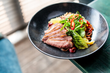 A gourmet dish featuring sliced medium-rare duck breast, garnished with fresh greens, red chili slices, and served with a side of grains and vegetables on a black plate.