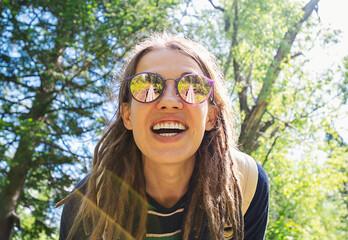 Smiling Woman with Long Dreadlocks and Sunglasses, Enjoying a Sunny Day in a Green Park Surrounded by Trees.